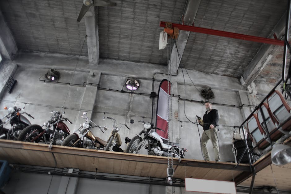 From below of bearded mechanic in casual outfit standing near motorcycles on special platform in garage and holding control panel in hand
