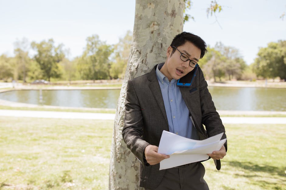 Asian businessman in corporate attire multitasking with phone and papers outdoors near a lake.